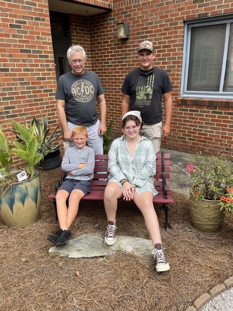 An adult and three children of varying ages sit on a bench in a flowerbed. 